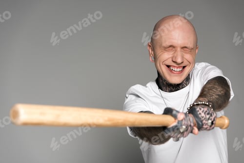 Preview: bald tattooed man with closed eyes hitting with baseball bat and laughing isolated on grey