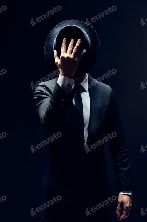 Preview: Man in suit hiding face behind his hat isolated on dark background