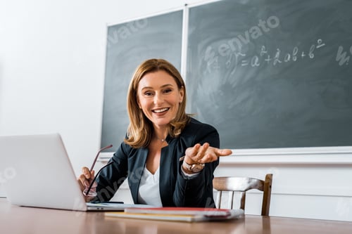 Preview: female teacher sitting at computer desk, holding glasses and looking at camera in classroom