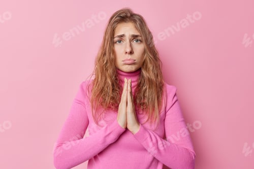 Preview: Upset good looking young woman with needy face begsfor something stands in pray pose looks sadly pur