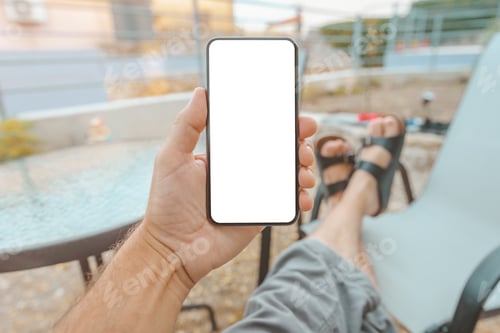 Preview: Man sitting on apartment terrace with feet on the table looking at mobile smart phone mockup screen