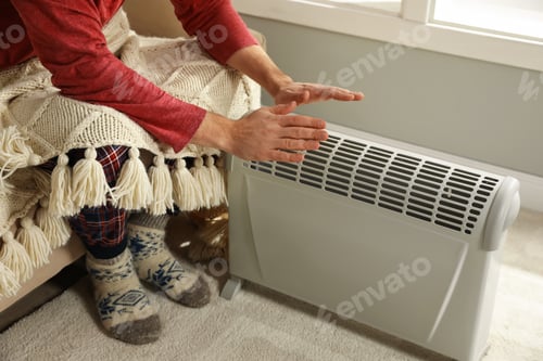 Preview: Man warming hands near electric heater at home, closeup