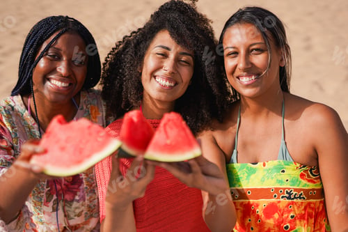 Preview: Young multiracial women smiling and holding fresh watermelon on camera at the beach