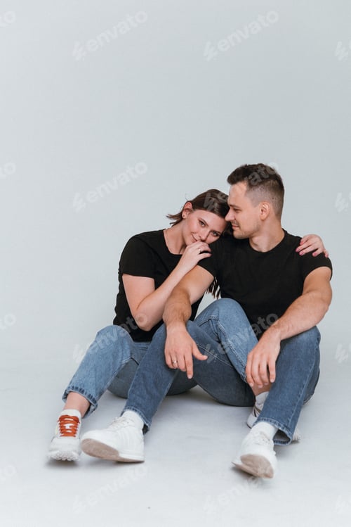 Preview: A young couple in love is sitting on a white background in black T-shirts and jeans hug and smile