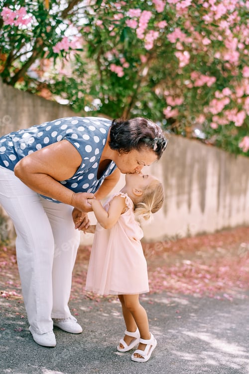 Preview: Granny kisses her little granddaughter leaning towards her near a flowering tree in the garden