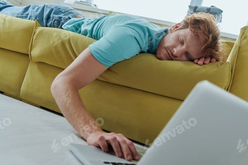 Preview: Young redhead man felt asleep while lying down on the couch and using laptop