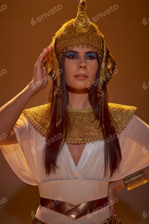 Preview: Portrait of brunette woman in egyptian outfit holding hand on hip on brown background with shadow
