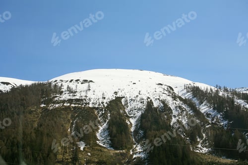 Preview: Snow-capped mountain under a clear blue sky, with sparse trees on the slopes.
