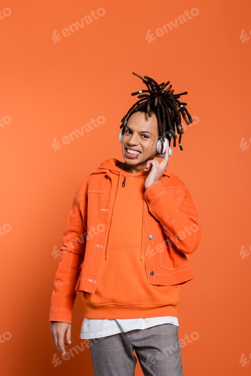 Preview: happy and multiracial man listening music in wireless headphones while looking at camera on coral