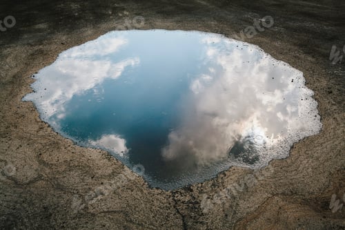 Preview: Rain drops falling onto a large puddle. A reflection of sky and clouds.