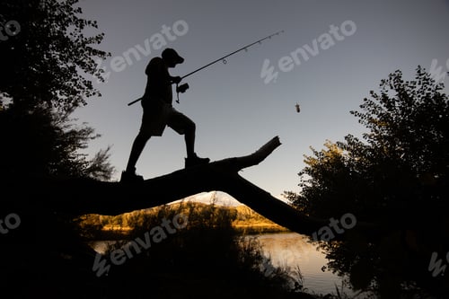 Preview: Low angle shot of a silhouette of a fisherman with a rod standing on a fallen tree log