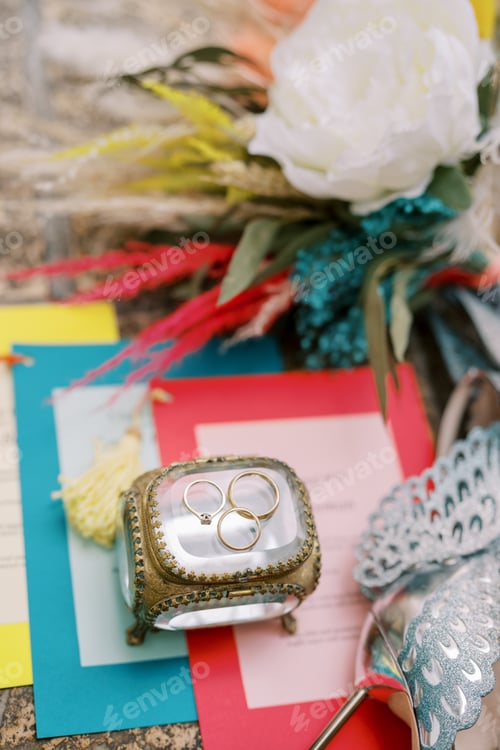Preview: Wedding rings lie on a glass box standing on the invitations next to the bouquet