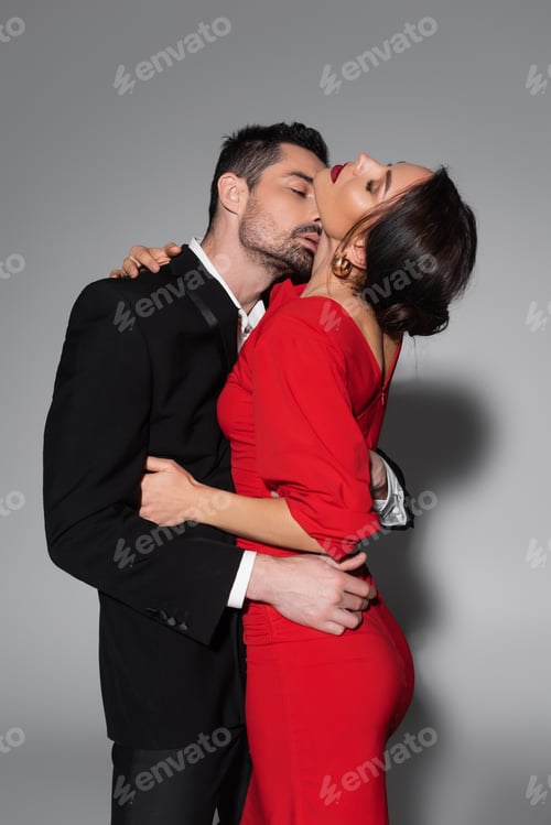Preview: Sexy man in suit kissing elegant woman in dress on grey background