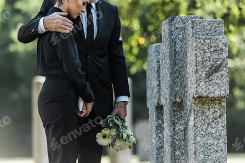 Preview: cropped view of senior man holding flowers while hugging woman near tombs