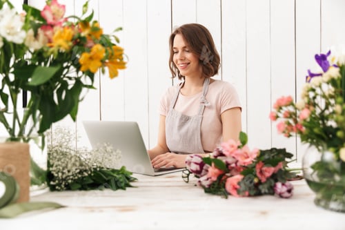 Preview: Cheerful florist lady standing with flowers using laptop computer.