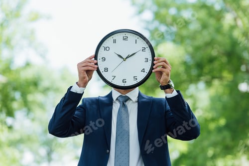 Preview: young man covering face with clock while standing in park