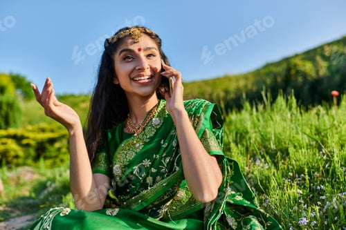 Preview: smiling young indian woman in green sari talking on smartphone while sitting on grass in summer