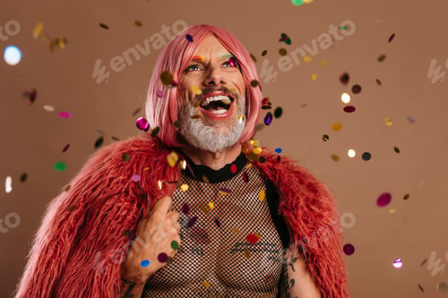Preview: Happy gay man in pink wig looking at the colorful confetti flying around against brown background