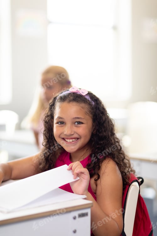 Preview: Portrait of smiling cute biracial elementary schoolgirl with book sitting at desk in classroom