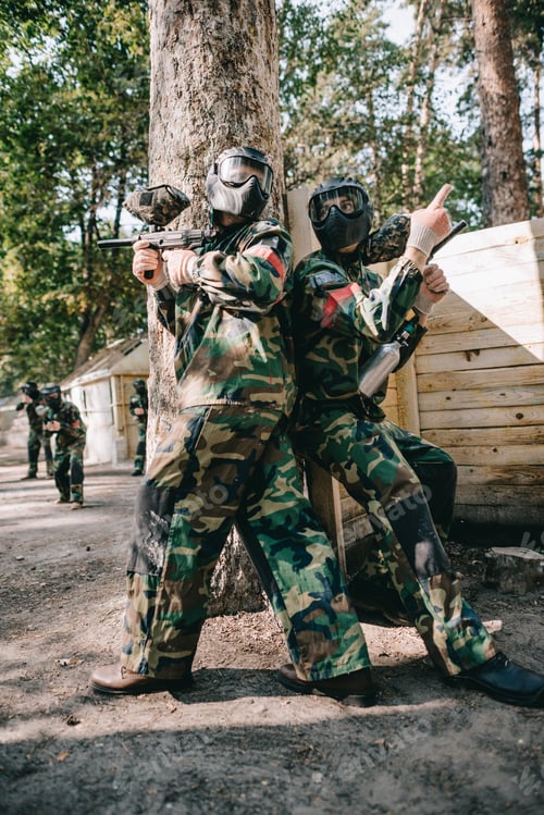 Preview: male paintball player in goggle mask and camouflage holding marker gun and pointing by finger to