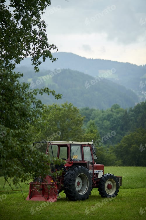 Preview: Vertical shot of a tractor in a green meadow with tree-covered mountains in the background