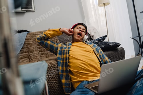 Preview: Teenage boy yawning while using laptop and sitting on floor at home