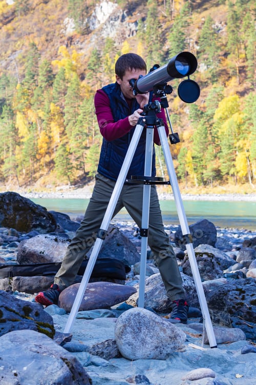 Preview: Full length shoit an attractive man in sportswear looking through telescope on bank of mountain rive