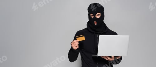 Preview: Hacker in balaclava holding laptop and credit card isolated on grey, banner