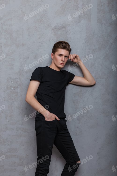 Preview: Model of a trendy young man in a stylish black t-shirt with a fashion hairstyle stands near a wall