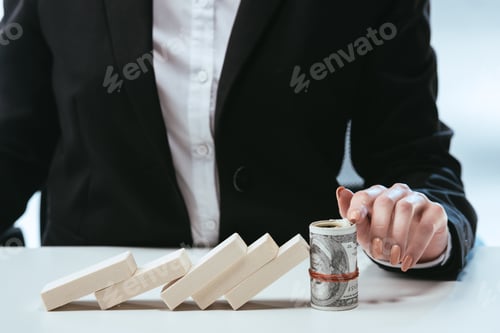 Preview: partial view of businesswoman sitting at table with fallen row of wooden blocks and money roll