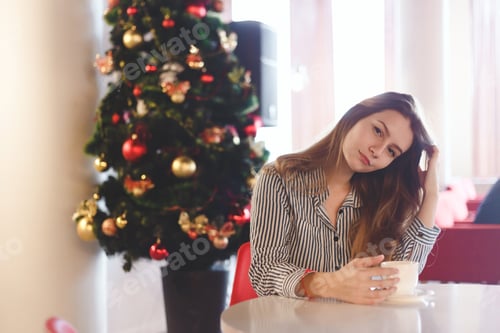 Preview: Girl drinking coffee sitting near a Christmas tree in cafe
