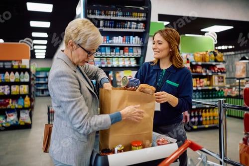 Preview: Happy cashier helping senior customer with packing groceries in paper bag at supermarket checkout.