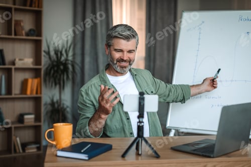 Preview: Smiling caucasian old man teacher with beard looks at smartphone and points at blackboard