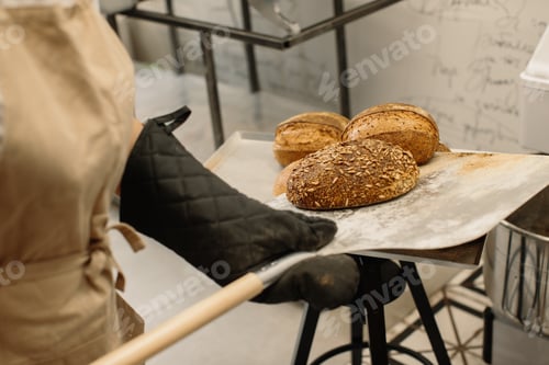 Preview: Female baker using a peel to take out a loaf of bread of the oven in a bakery