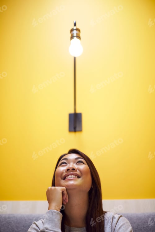 Preview: Woman Sitting Under Light Bulb In Office Suggesting Inspiration Or Idea