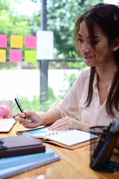 Preview: Smiling young woman making note on notebook while sitting at her office desk.