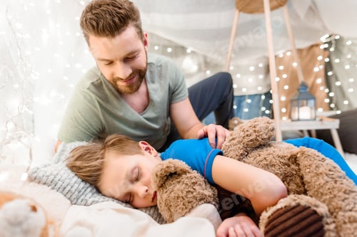 Preview: Smiling father looking at cute little son sleeping with teddy bear