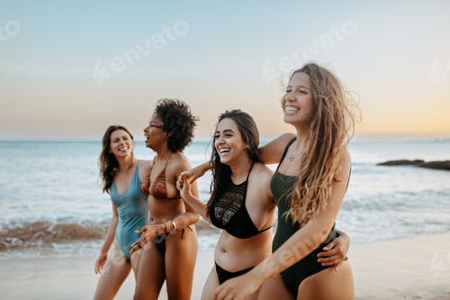 Preview: Joyful girlfriends in swimwear walking on beach at sunset and enjoying free time together, laughing