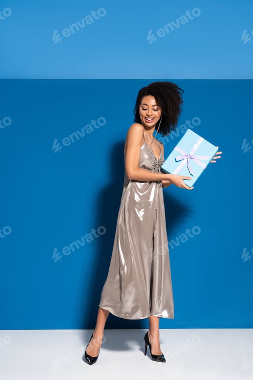 Preview: happy beautiful african american woman in silver dress holding gift on blue background