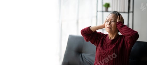 Preview: Asian mature woman sitting alone at home touching head feeling headache migraine.