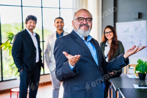 Preview: Confident oldIndian business leader standing with folded hands, colleagues out of focus in office