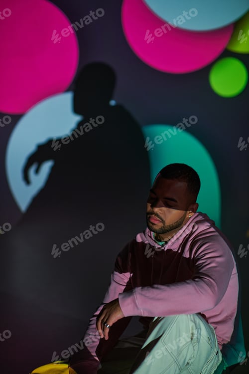 Preview: good looking man in pink hoodie sitting on floor in digital projector lights, fashion concept