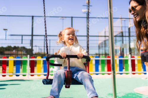 Preview: Happy baby girl swinging at playground with mother watching