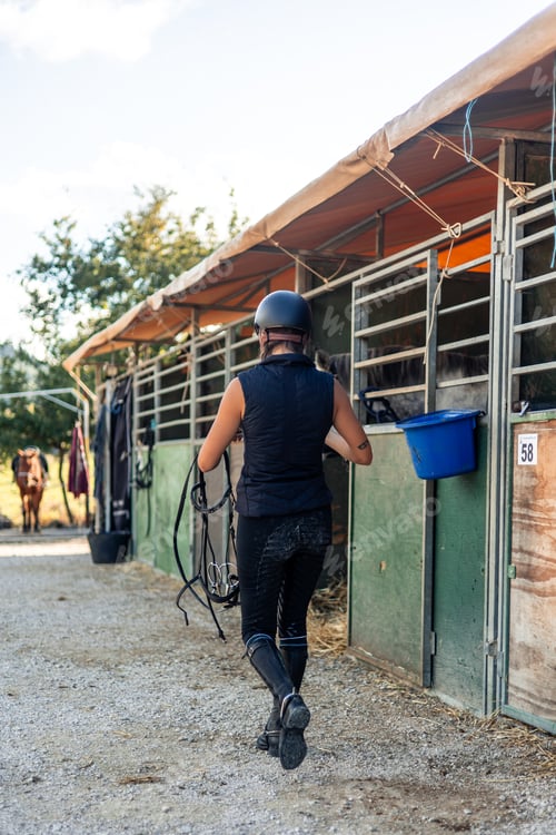 Preview: vertical Female rider with a helmet walking through the stables carrying saddles at the ranch