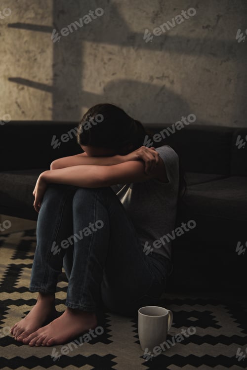 Preview: emotional crying woman hugging her knees while sitting on floor at sofa