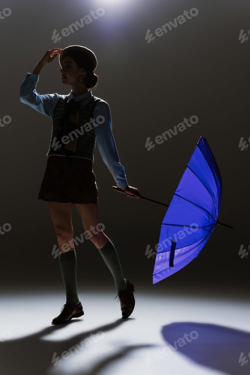 Preview: silhouette of elegant young woman in beret and skirt posing with blue umbrella on grey