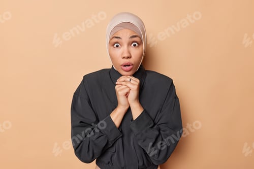 Preview: Indoor shot of shocked concerned woman keeps hands under chin gasps startled keeps mouth opened