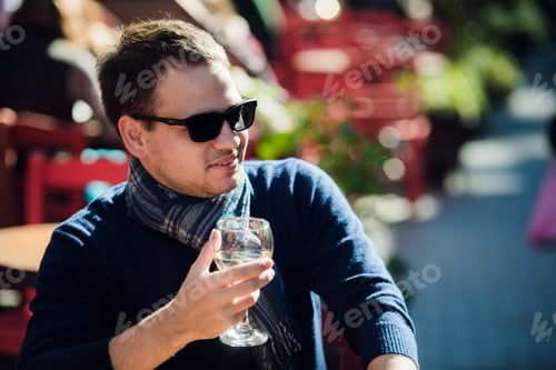 Preview: Young man sitting at outdoor bar table having a glass of white wine and looking to camera