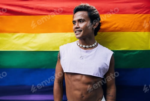 Preview: Smiling young gay man in stylish clothes standing against pride flag