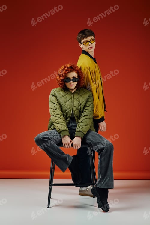 Preview: beautiful woman in sunglasses sitting on chair next to her fashionable boyfriend on red backdrop